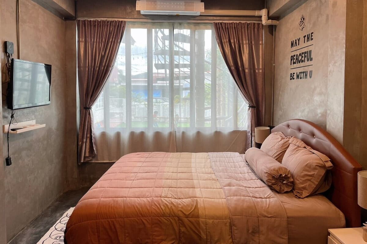 Un dormitorio con cama, televisor, ventana con cortinas y una frase motivacional en la pared.