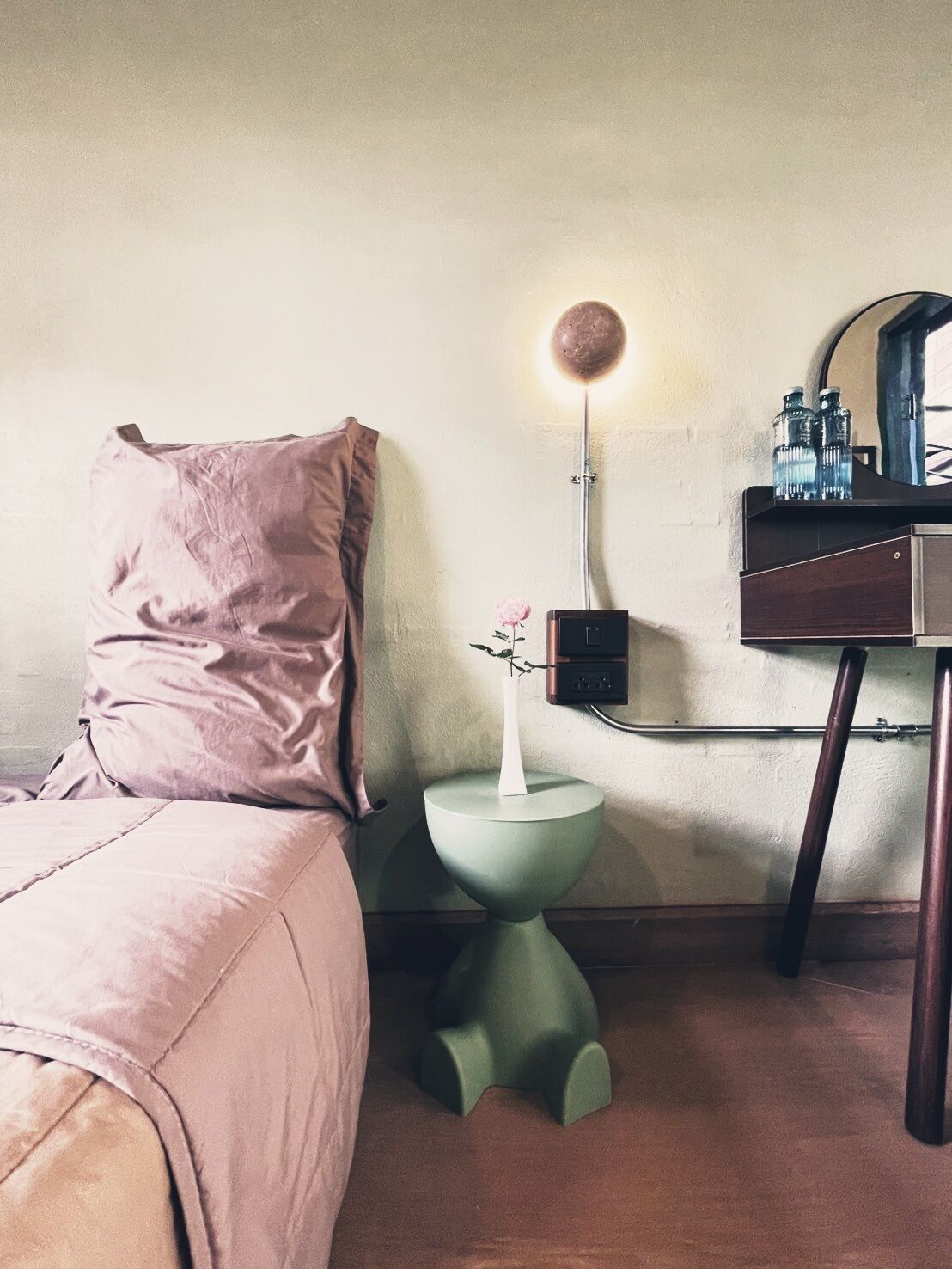 A hotel room with a bed, a green nightstand with a vase and a lamp, and a wooden dresser with bottles.