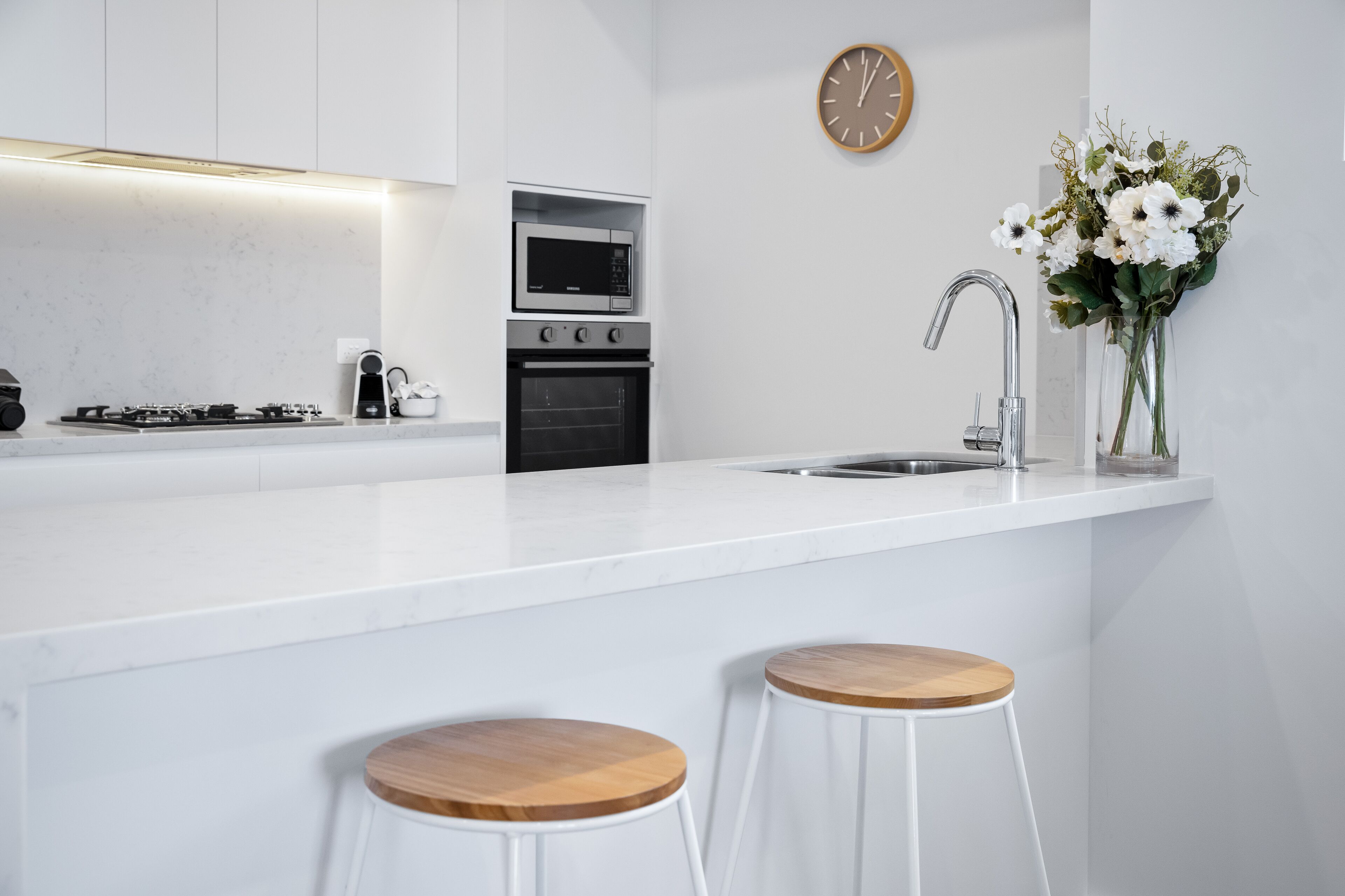 A modern kitchen with white countertops, a built-in oven, a microwave, a sink, and two wooden stools.