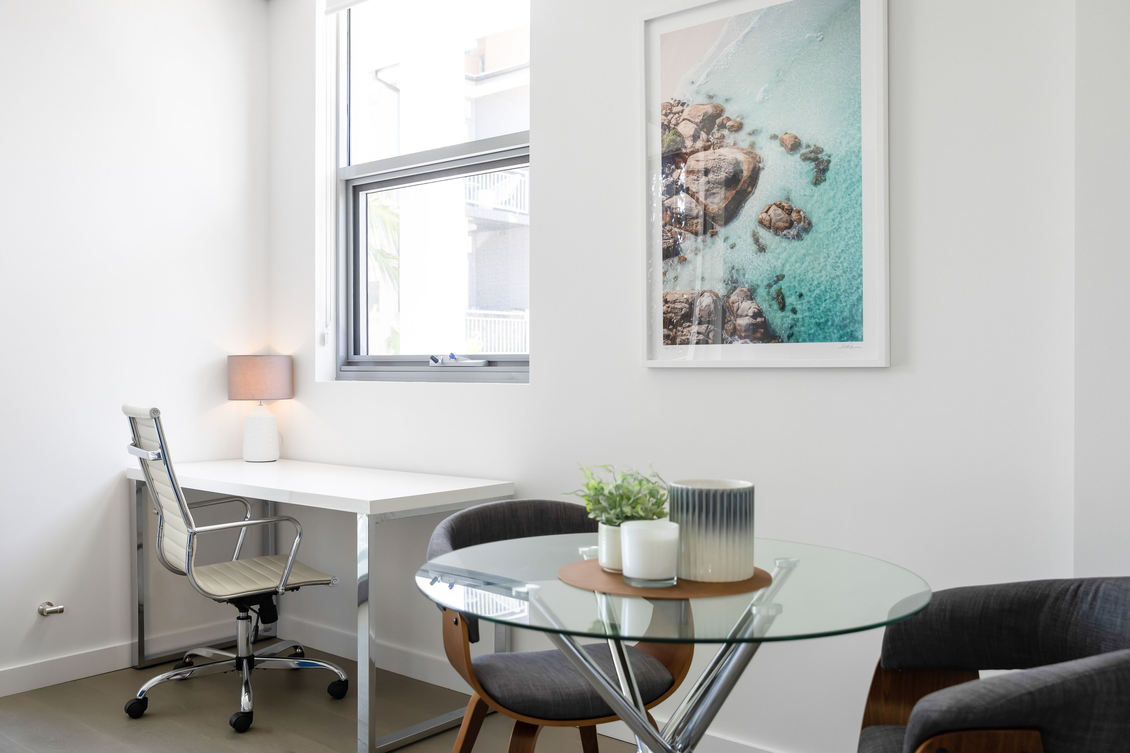 A modern room with a white desk, a glass table, and two chairs. There is a window with a view of buildings and a framed artwork on the wall.
