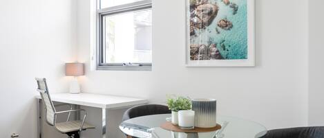 A modern room with a white desk, a glass table, and two chairs. There is a window with a view of buildings and a framed artwork on the wall.