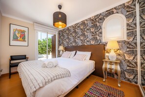 Ein Schlafzimmer mit einem großen Bett, einem Nachttisch mit Lampe, einem Sessel, einem gerahmten Kunstwerk und einer Wand mit Muster.