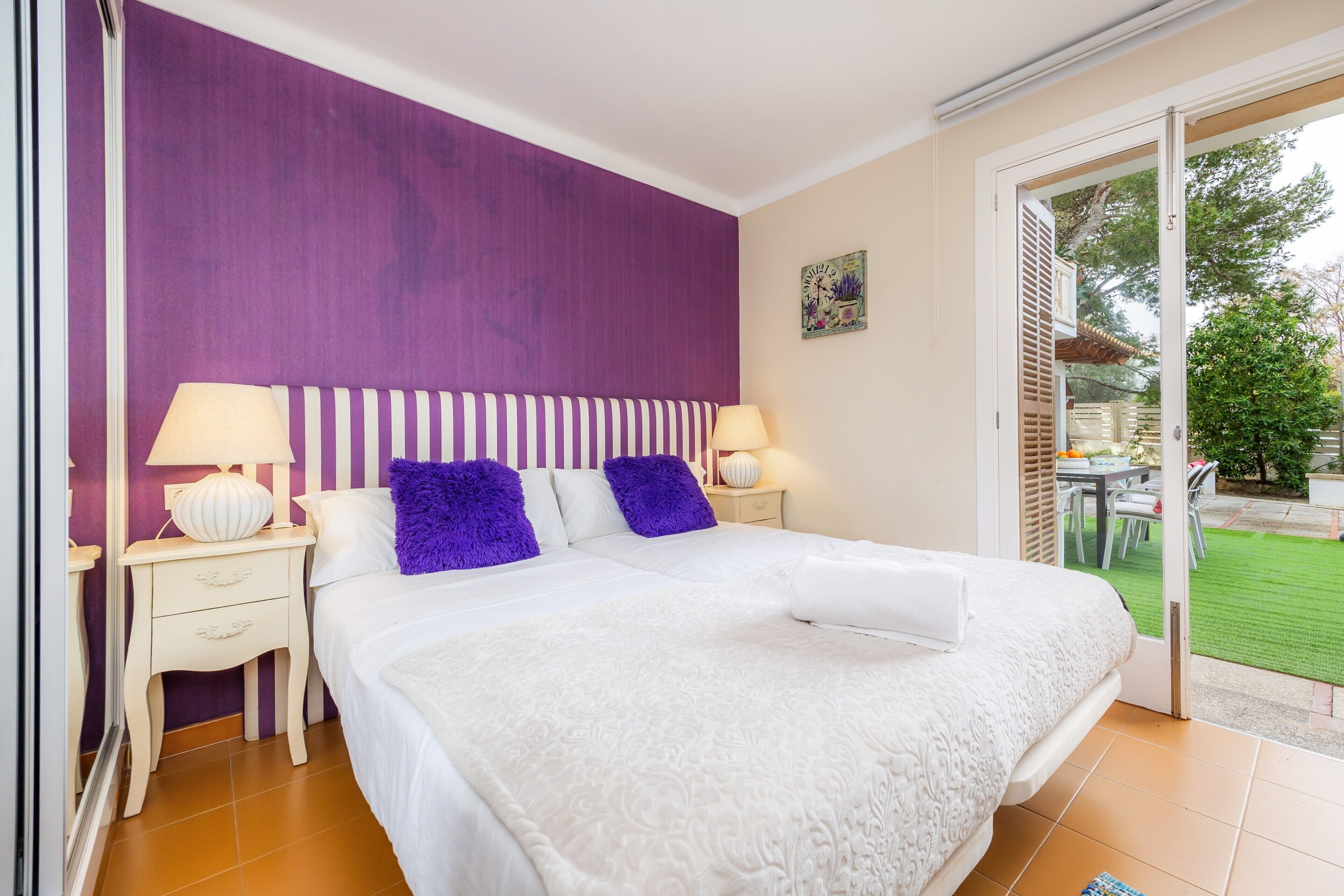 Un dormitorio con una cama grande, dos mesitas de noche, un cabecero morado y vistas a un jardín a través de una puerta abierta.