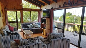 A cozy living room with striped sofas, a small table, and a bookshelf. A dog is visible on the balcony.