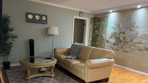 A living room with a beige leather sofa, a glass coffee table, a patterned rug, a wall clock, a lamp, a potted plant, and a mural. - Cozy Condo in Downtown Long Beach, 2 min walk to the Beach,parking place+garage  (Long Beach)