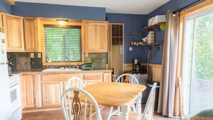 A kitchen with wooden cabinets, a granite countertop, a sink, and a dining area with a wooden table and white chairs. - Cozy Riverfront Cottage (Hersey)