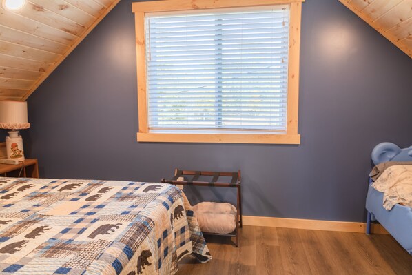 A bedroom with a sloped ceiling, a window with blinds, a bed with a patterned quilt, a small chair, and a blue bench.