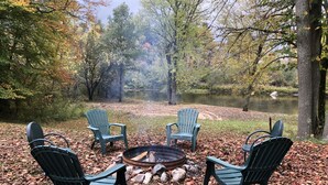 A circular seating area with four Adirondack chairs around a fire pit, set on a ground covered with fallen leaves, with a lake and trees in the background. - Cozy Riverfront Cottage (Hersey)