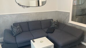 A grey sectional sofa with patterned cushions, a white side table with a remote control and a decorative item, and a mirror with a decorative frame on the wall.