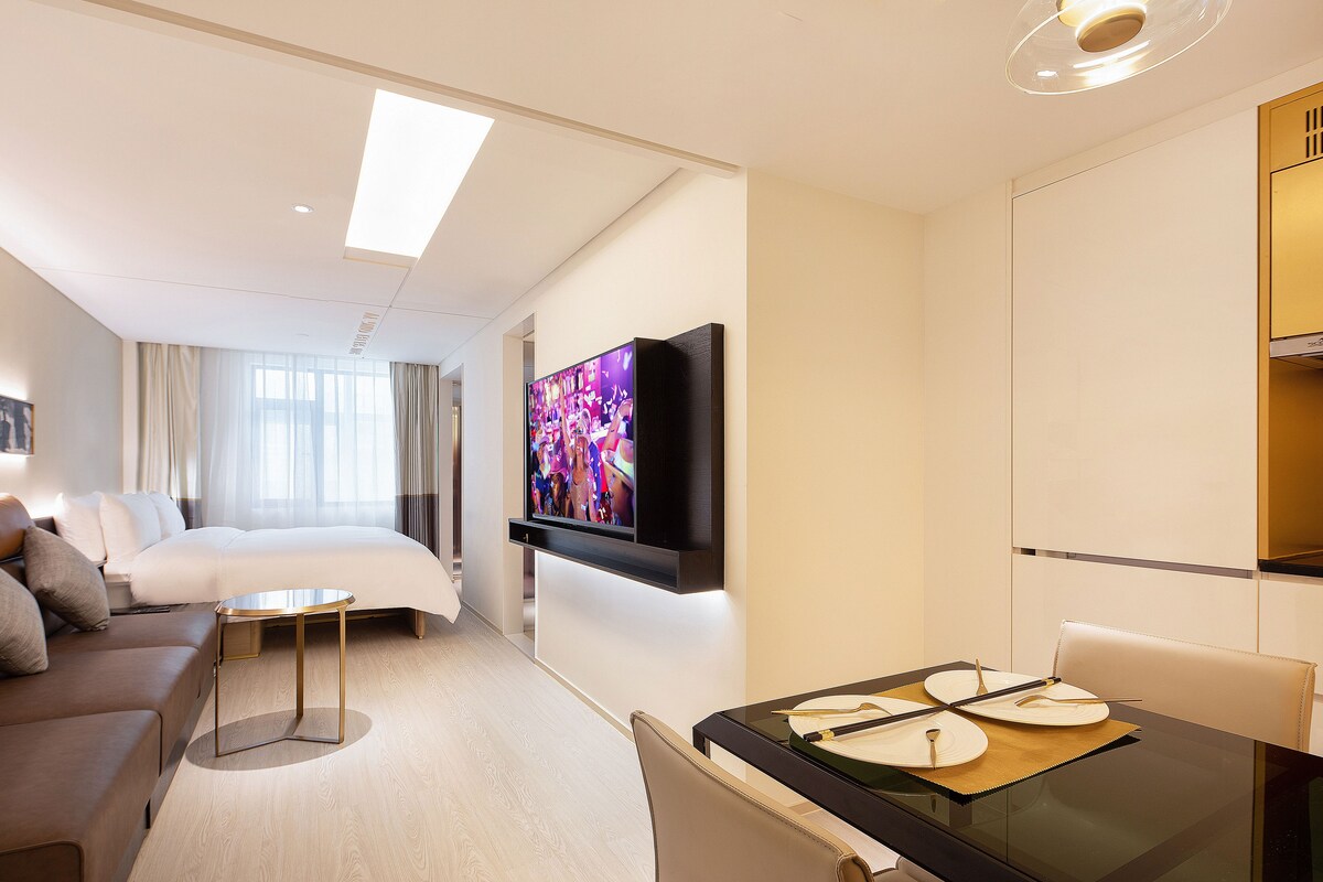 A hotel room with a bed, a TV mounted on the wall, a dining table with a placemat, and a sofa.
