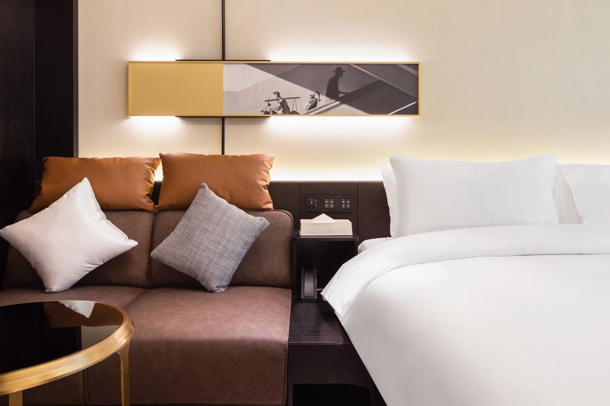 Ein Hotelzimmer mit Bett, einer Couch mit Kissen, einem kleinen Tisch, einem Nachttisch mit Lampe und einem an der Wand hängenden Kunstwerk.