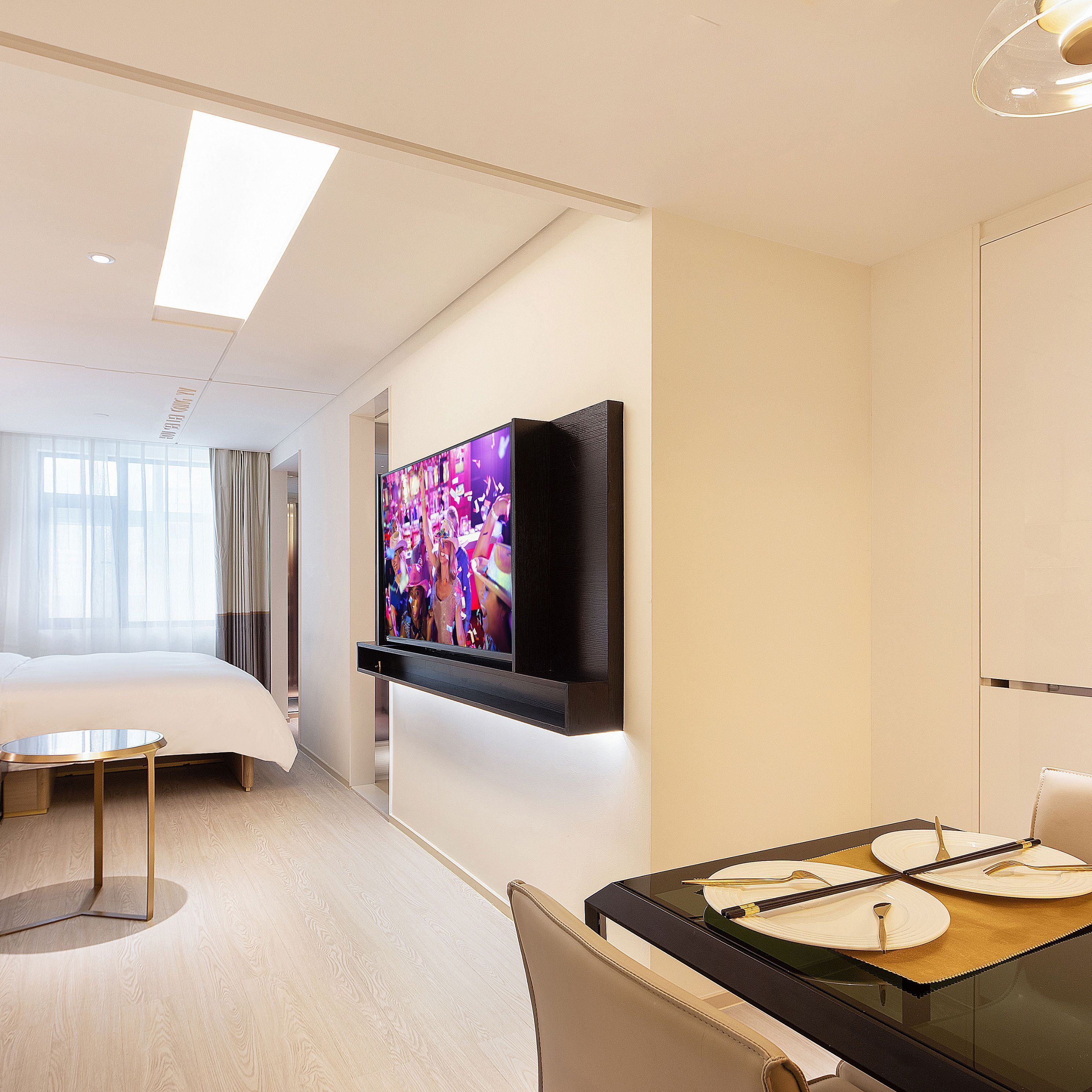 Una habitación de hotel moderna con un televisor de pantalla plana grande montado en la pared, una cama con cabecera blanca, una mesa de centro redonda de vidrio y una zona de comedor con una mesa preparada para cuatro.