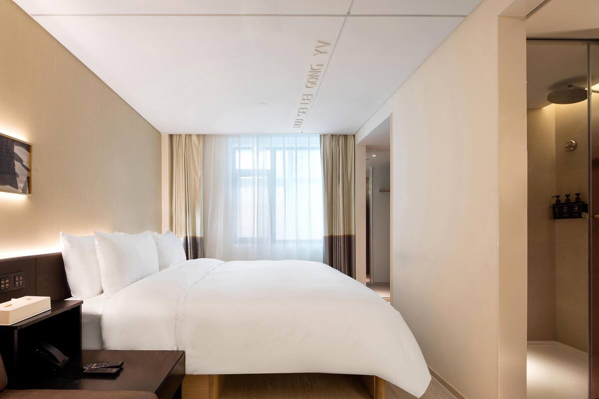 Ein Hotelzimmer mit einem großen Bett, einem Nachttisch mit Fernbedienung, einem Fenster mit Vorhängen und einem Duschbereich.