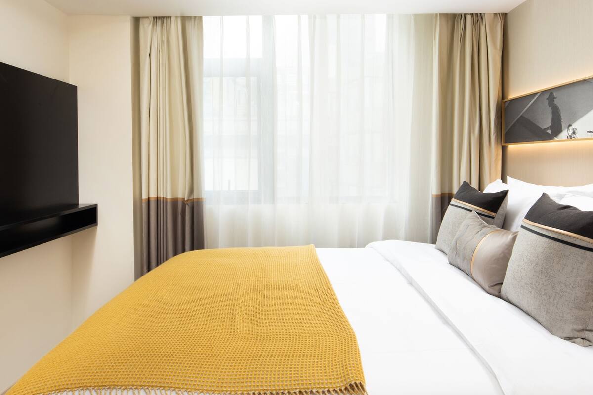 Ein Hotelzimmer mit einem großen Bett, einem flachbildfernseher an der Wand und einem Fenster mit durchsichtigen Vorhängen.
