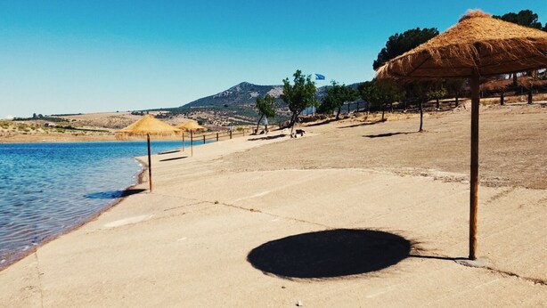 Una playa con un paseo marítimo de concreto, una sombrilla de paja y una montaña al fondo.