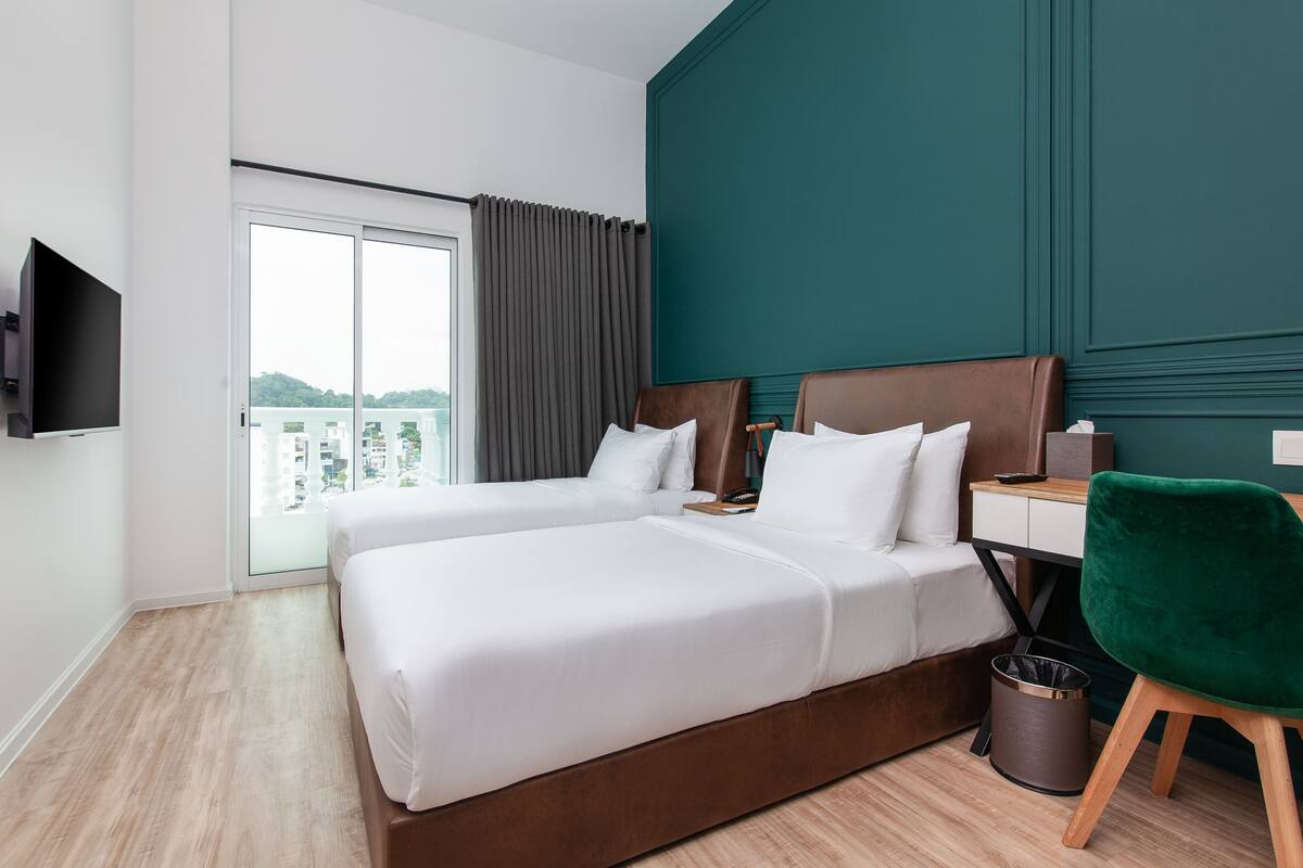 A hotel room with two beds, a desk, a TV, a green chair, and a view of the city.