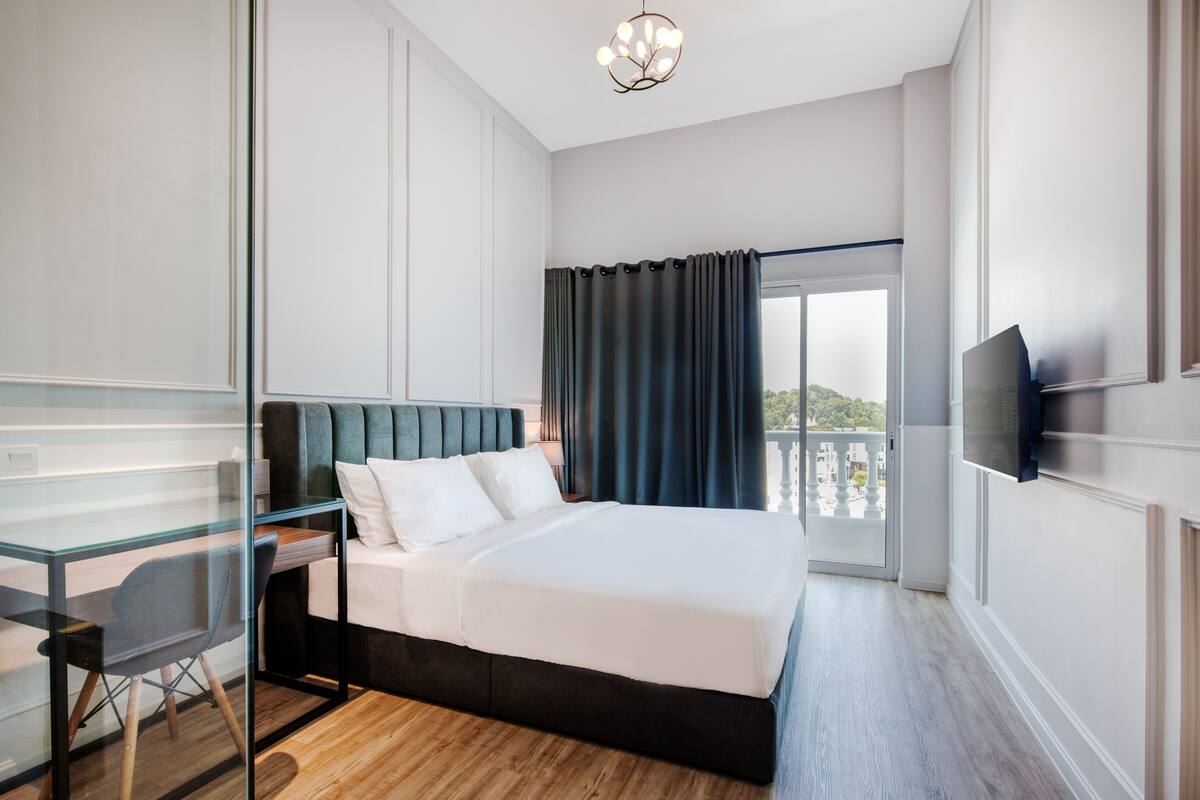 A modern hotel room with a large bed, a desk with a chair, a TV mounted on the wall, and a balcony with a view.