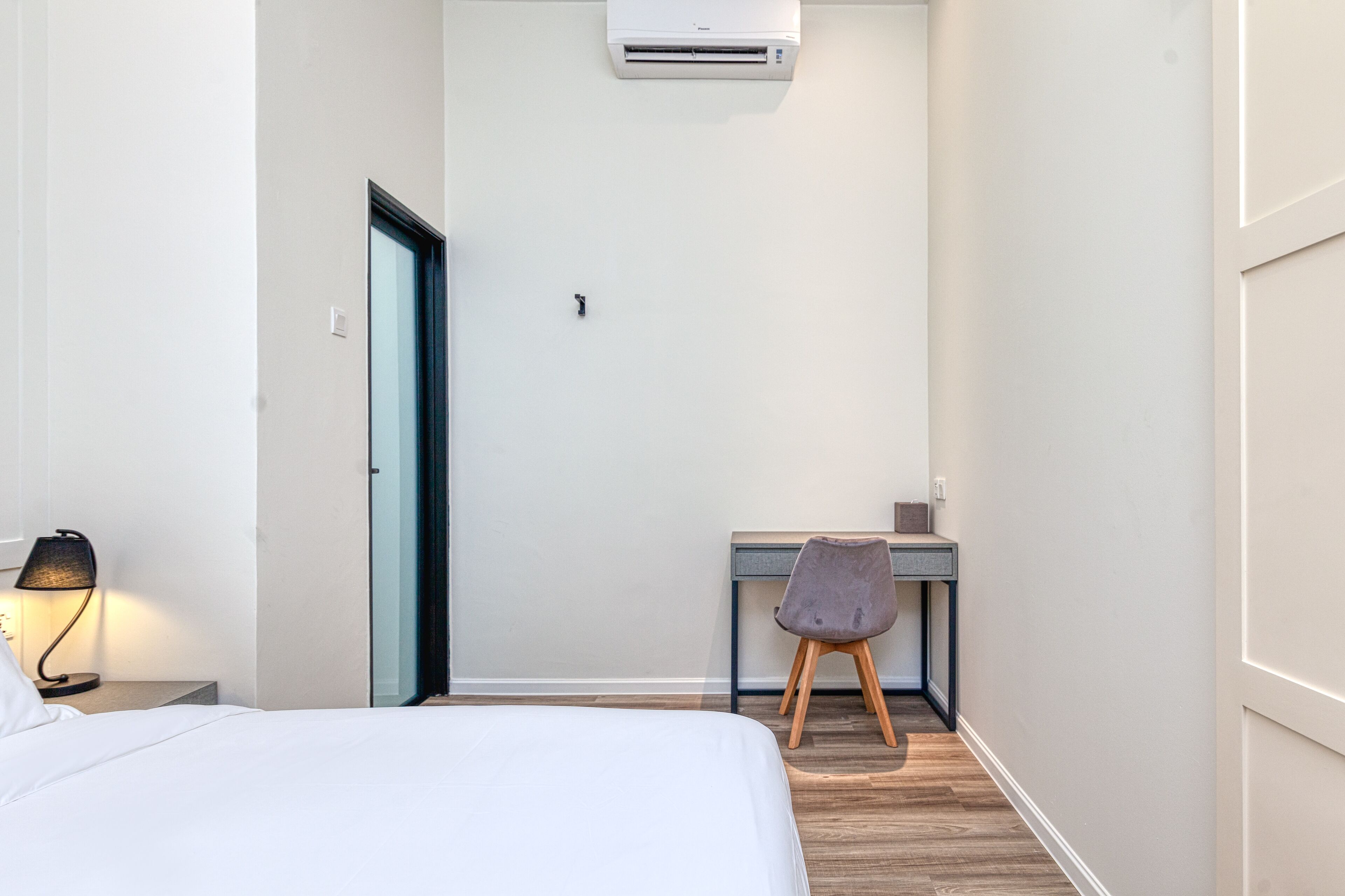 A hotel room with a bed, a desk, a chair, an air conditioner, and a nightstand with a lamp.