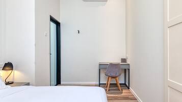 A hotel room with a bed, a desk, a chair, an air conditioner, and a nightstand with a lamp.