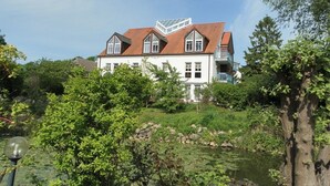 A large white building with a red roof, surrounded by greenery and a pond. - Haus am Teich, Whg. D16 (Ostseebad Boltenhagen)