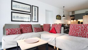 A modern living room with a grey sofa set, red patterned cushions, and a wooden coffee table. There are framed cityscape pictures on the wall. - Haus am Teich, Whg. D16 (Ostseebad Boltenhagen)