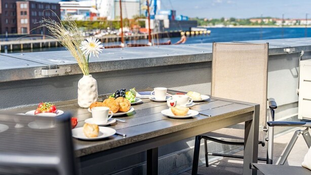 Una mesa de comedor con platos de comida y bebidas, un jarrón con flores, y vista al puerto con edificios y grúas al fondo.