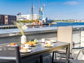 A dining table with plates of food and drinks, a vase with flowers, and a view of the harbor with buildings and cranes in the background. - Hafenspitze App. 09, Deutschland (Wismar)