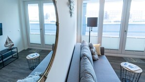 Una sala de estar moderna con un espejo grande, un sofá gris y vistas al agua a través de amplios ventanales.