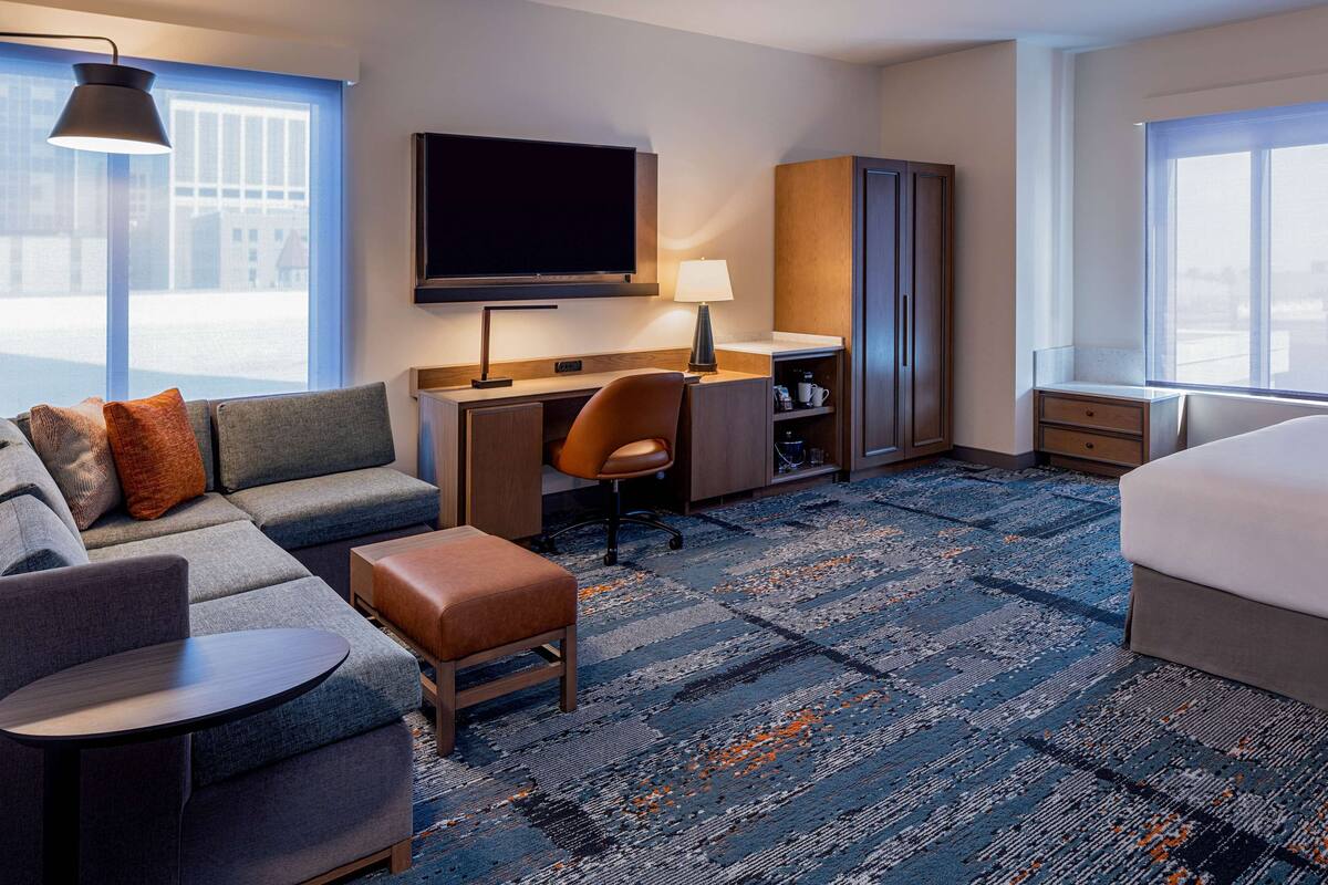 Ein Hotelzimmer mit Sofa, Schreibtisch, Stuhl, Fernseher, Kleiderschrank und Bett.