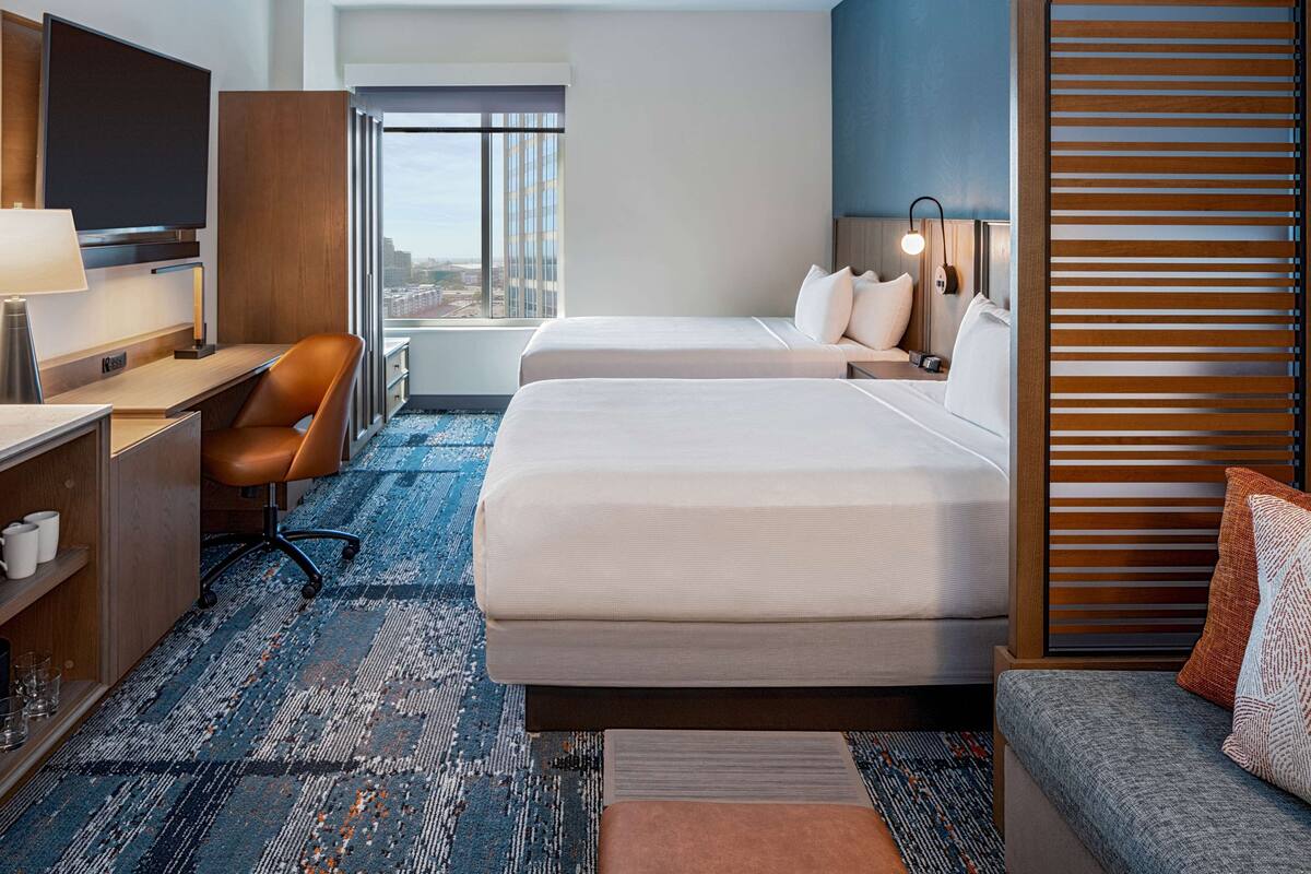 Ein Hotelzimmer mit zwei Betten, einem Schreibtisch, einem Stuhl, einem Fernseher und Blick auf die Stadt.