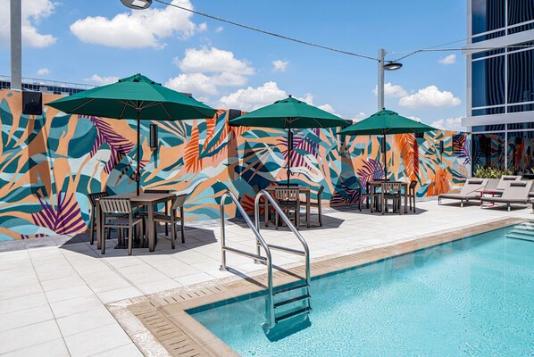 A poolside area with a mural, lounge chairs, and tables under green umbrellas.