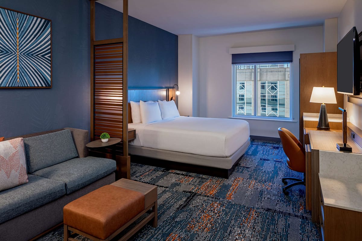 Ein Hotelzimmer mit Bett, Sofa, Schreibtisch, Fernseher und einem Fenster mit Jalousien.