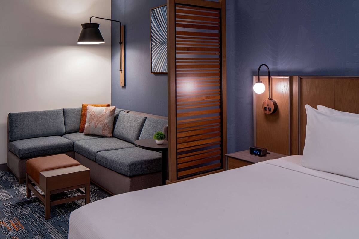 Ein modernes Hotelzimmer mit Ecksofa, Holzabtrennung, Nachttisch mit Lampe und Bett mit weißen Bettwäsche.