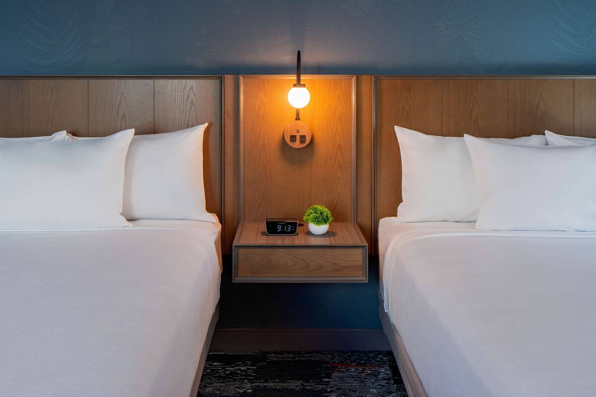 Ein Hotelzimmer mit zwei Betten, einem Nachttisch mit einer Uhr und einer kleinen Pflanze sowie einer wandmontierten Lampe.