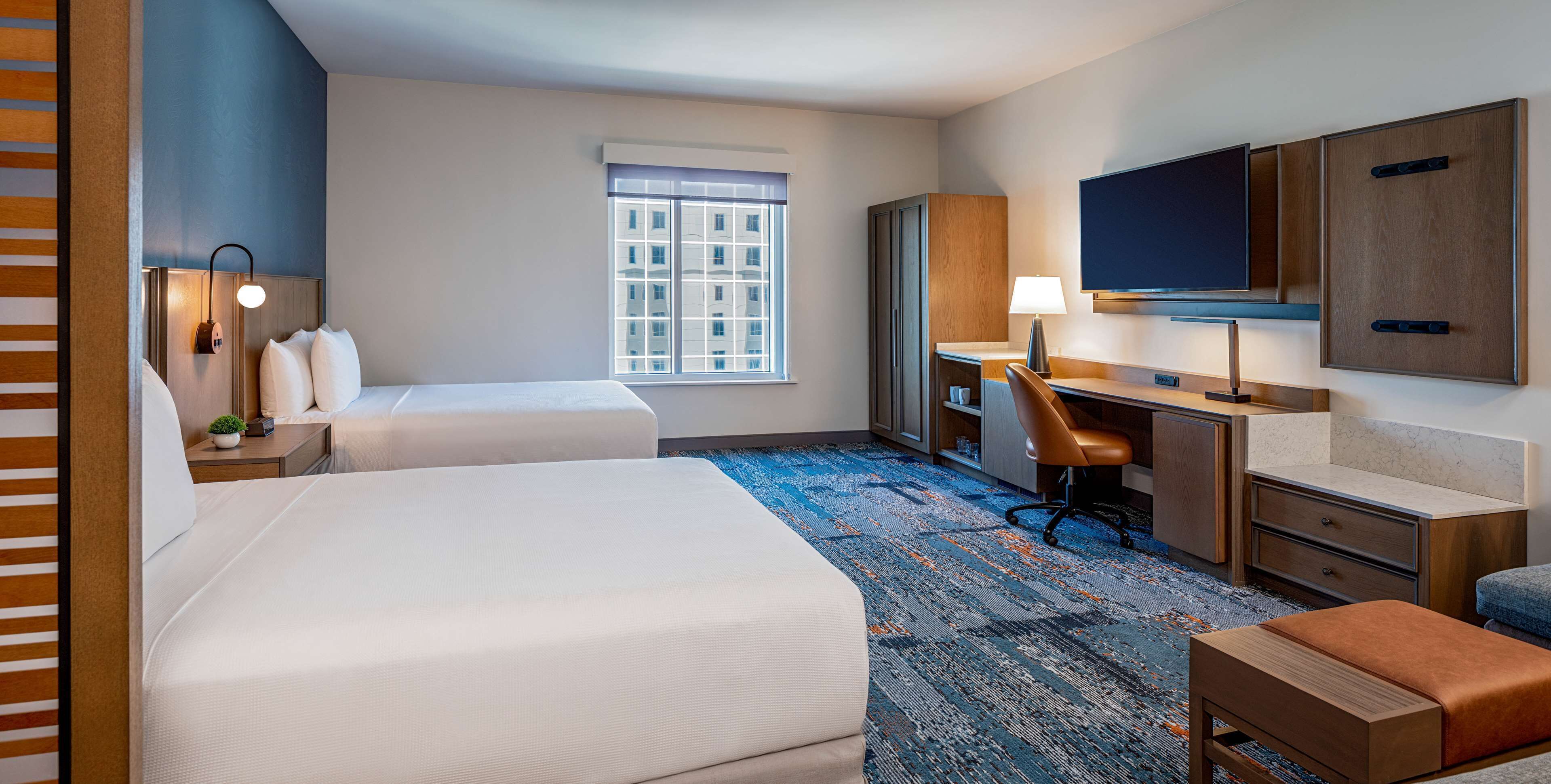 A hotel room with two beds, a desk, a chair, a TV, and a window with blinds.
