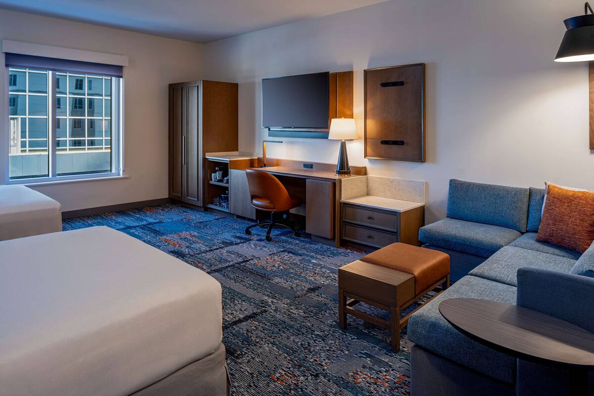 Ein Hotelzimmer mit Bett, Schreibtisch, Sessel, Sofa, einem kleinen Tisch, einer Lampe und einem Fernseher.