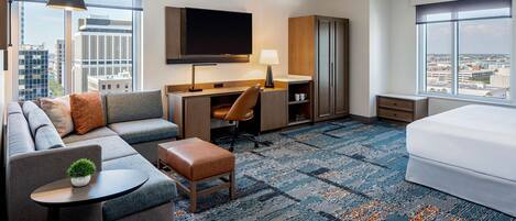 Quarto de hotel com sofá, escrivaninha, televisão, candeeiro, guarda-roupa e cama.