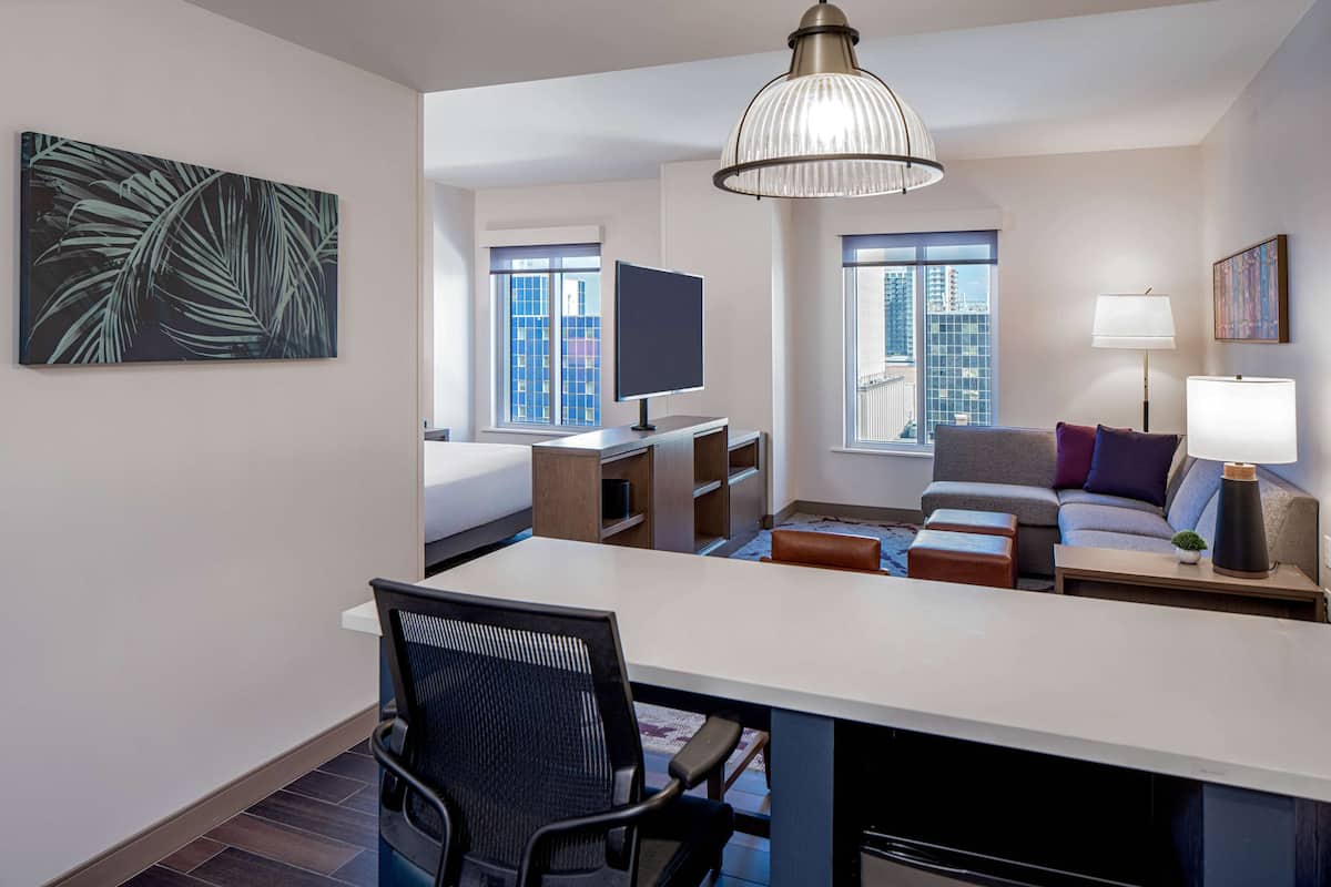 A modern hotel room with a large window, a flat-screen TV, a desk with a chair, a sofa, and a lamp.
