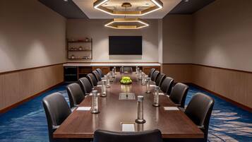 A conference room with a long table, chairs, a projector screen, and a shelf with books and decorative items.