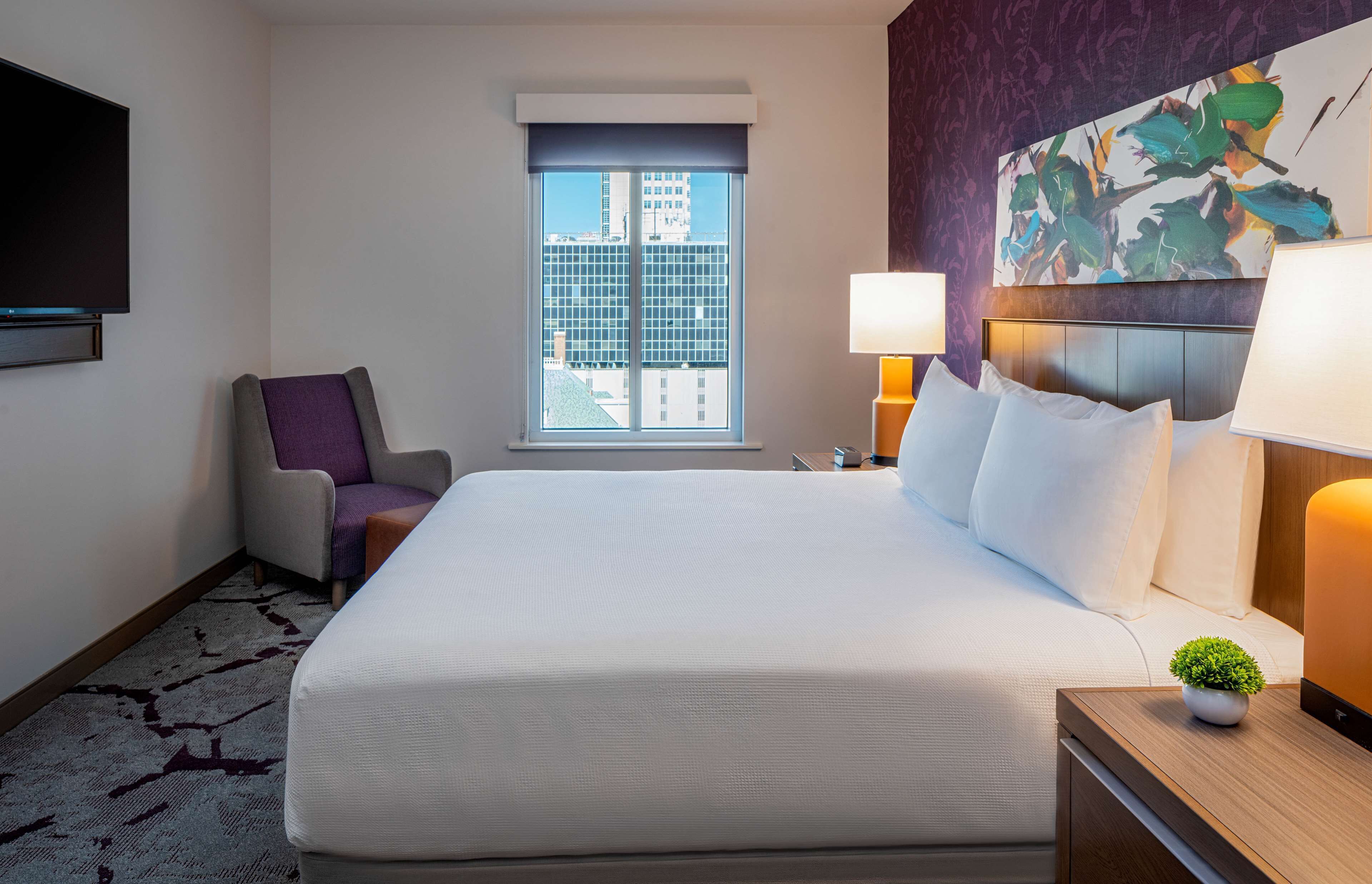 A hotel room with a large bed, a chair, a TV, a window with a city view, and a painting on the wall.