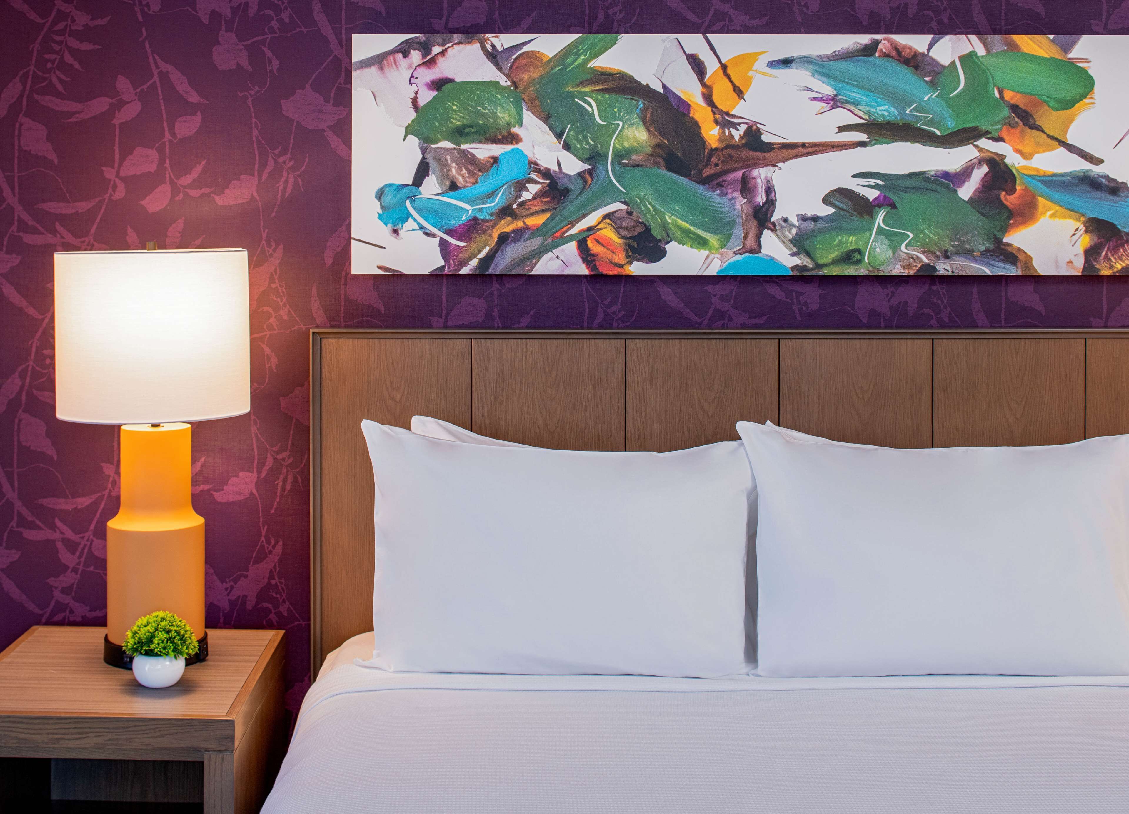 Ein Hotelzimmer mit einem hölzernen Kopfteil, einem weißen Bett mit zwei Kissen, einem Nachttisch mit Lampe und einer kleinen Pflanze sowie einem farbenfrohen, abstrakten Gemälde an der Wand.