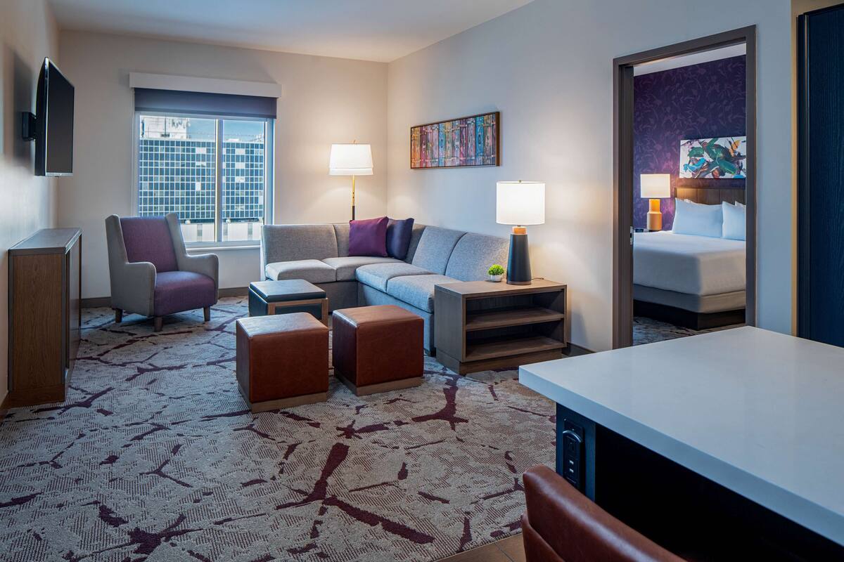 A hotel room with a sofa, armchairs, a dining table, a TV, and a bed with a headboard.