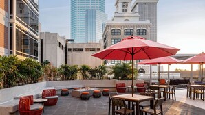 Un patio al aire libre con mobiliario y un hogar para fuego, rodeado de plantas y vistas a un paisaje urbano con edificios modernos.
