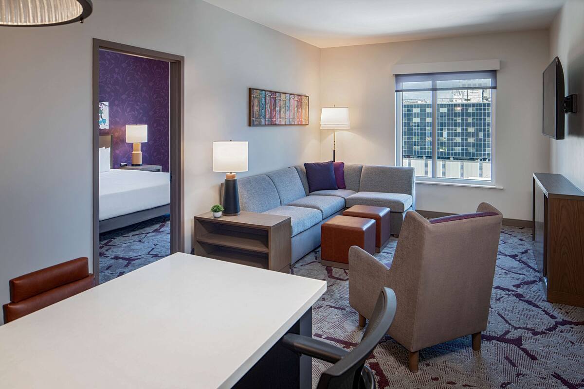 A hotel room with a bed, a sofa, a dining table, a TV, and a window with a city view.