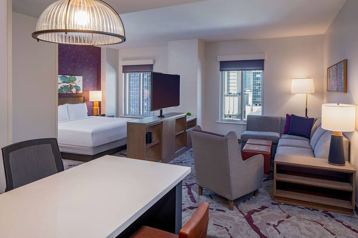 A hotel room with a bed, desk, TV, sofa, and windows with city views.