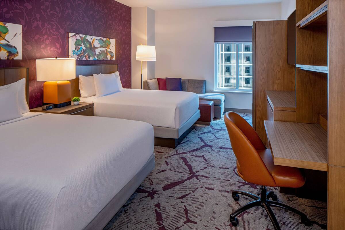 A hotel room with two beds, a desk, a chair, a lamp, and a window with a view of buildings.