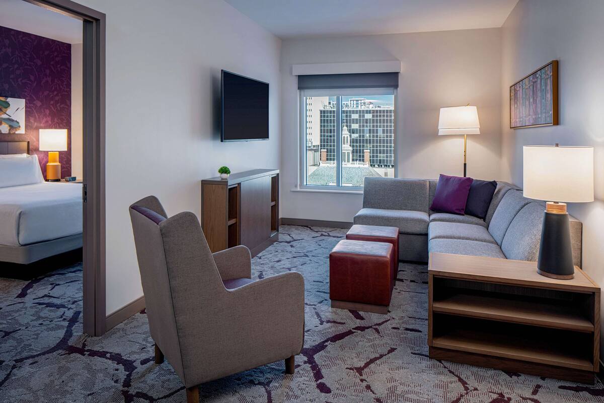 A hotel room with a bed, a sofa, a chair, a TV, and a window with a city view.