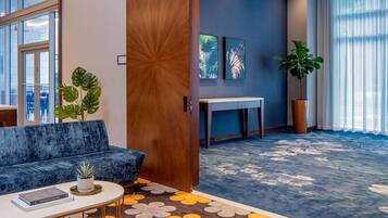 A modern lobby with a blue sofa, a coffee table with a book, a large potted plant, and a patterned carpet.