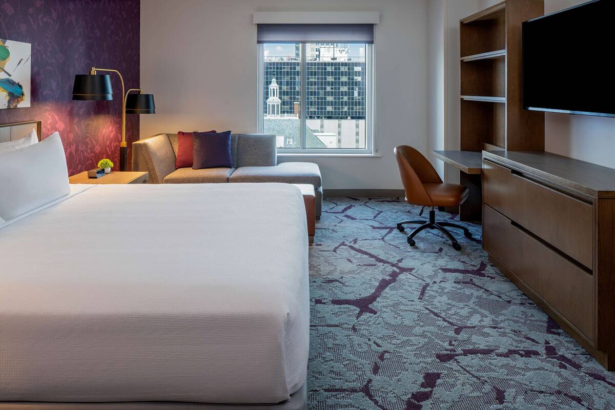 A hotel room with a large bed, two sofas, a desk, a TV, and a window with a city view.
