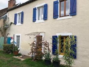 Un edificio de dos pisos con contraventanas azules, un pequeño jardín con plantas en macetas y una puerta visible.